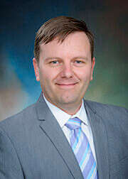 Professional portrait of UTMB Health professional, Alejandro Villasante-Tezanos, PhD, wearing a gray suit and light blue striped tie against a blue gradient background.